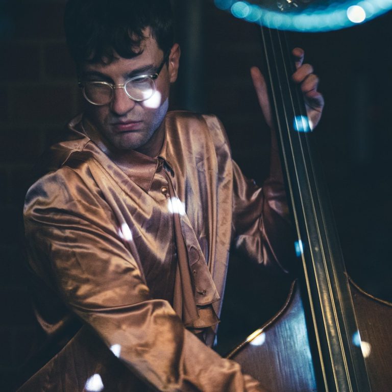 Lukas Growe am 5-Saiter Kontrabass blickt zur Seite beim spielen, dabei trägt er ein lachsfarbenes glänzendes Hemd
